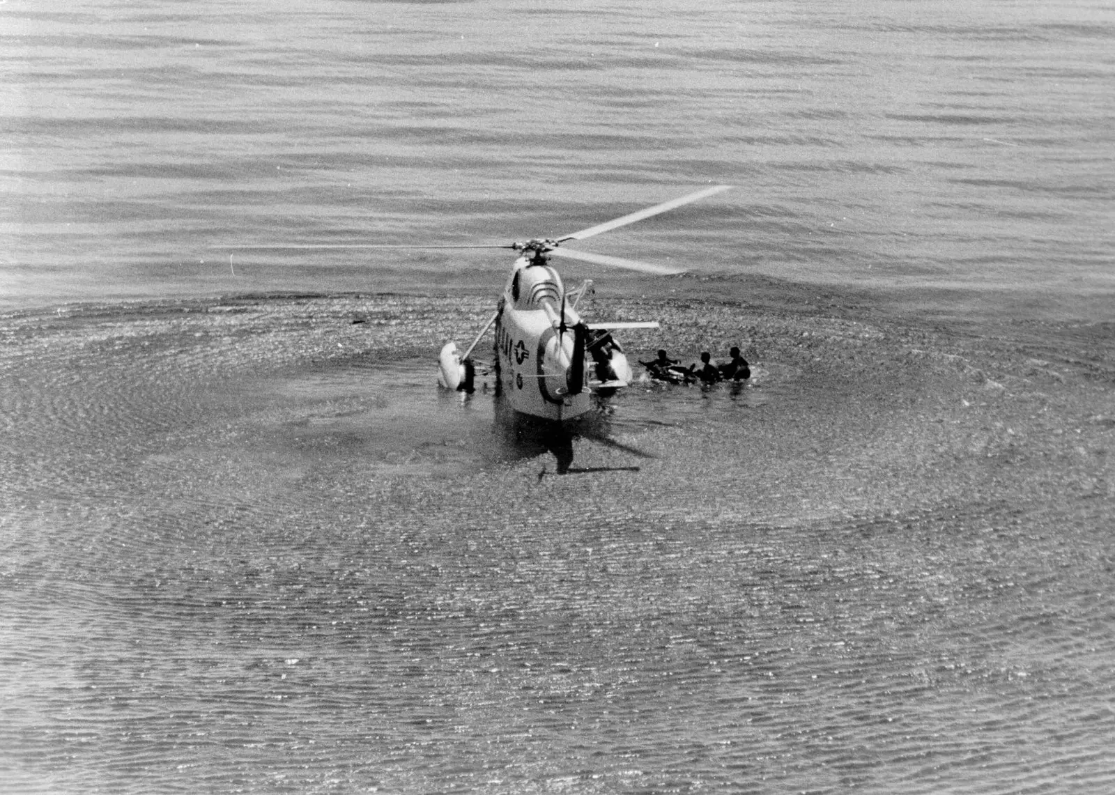 Foto en blanco y negro de 1980: un helicóptero HH-52 de la Guardia Costera de Estados Unidos posado sobre el mar en calma, con el rotor girando y las ondas que levanta a su alrededor. A su derecha, varias personas en el agua, que se ven como siluetas.